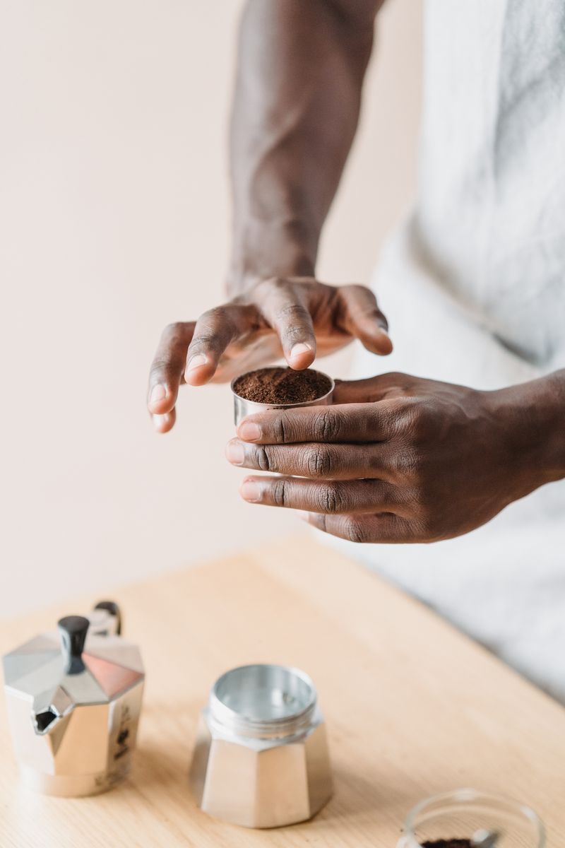 Hands measuring coffee grounds into a moka pot filter in a home kitchen