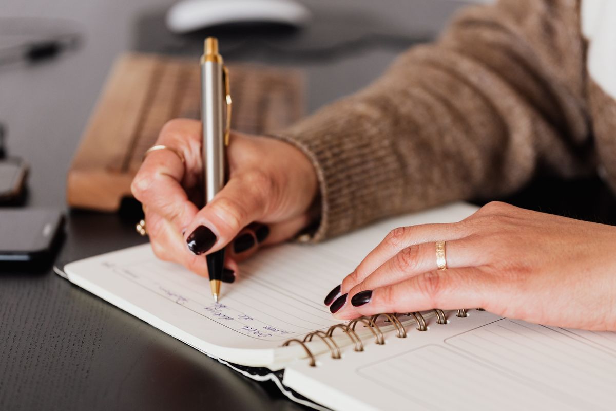 Person writing in a spiral notebook at a desk beside a keyboard and phone