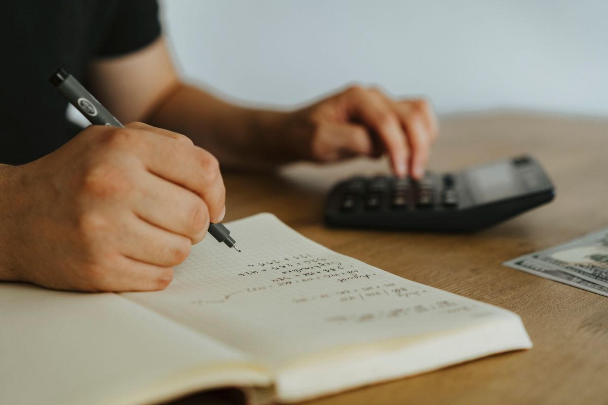 Hands writing calculations in a notebook beside a calculator on a wooden table