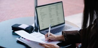 Person writing on paper at a table beside an open laptop