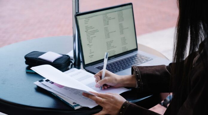Person writing on paper at a table beside an open laptop