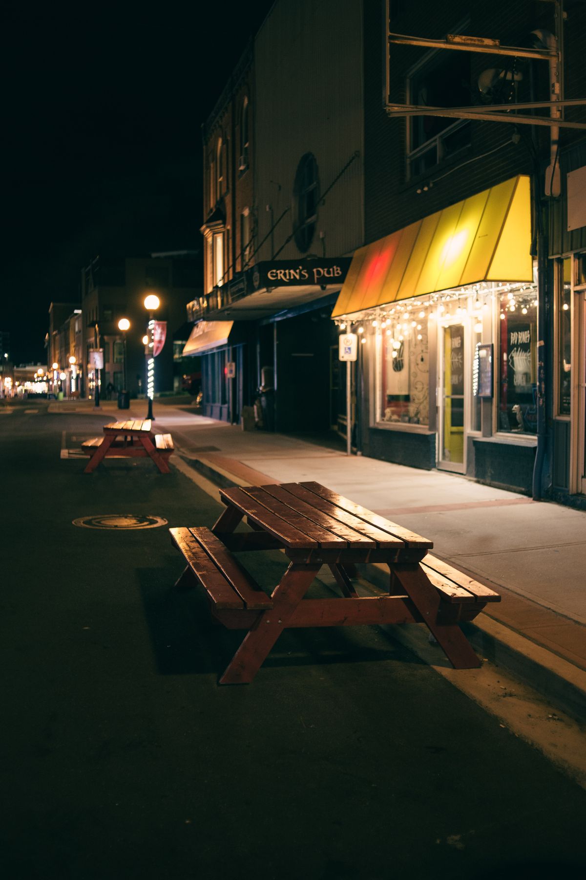 Wooden outdoor seating beside illuminated shops on a street at night